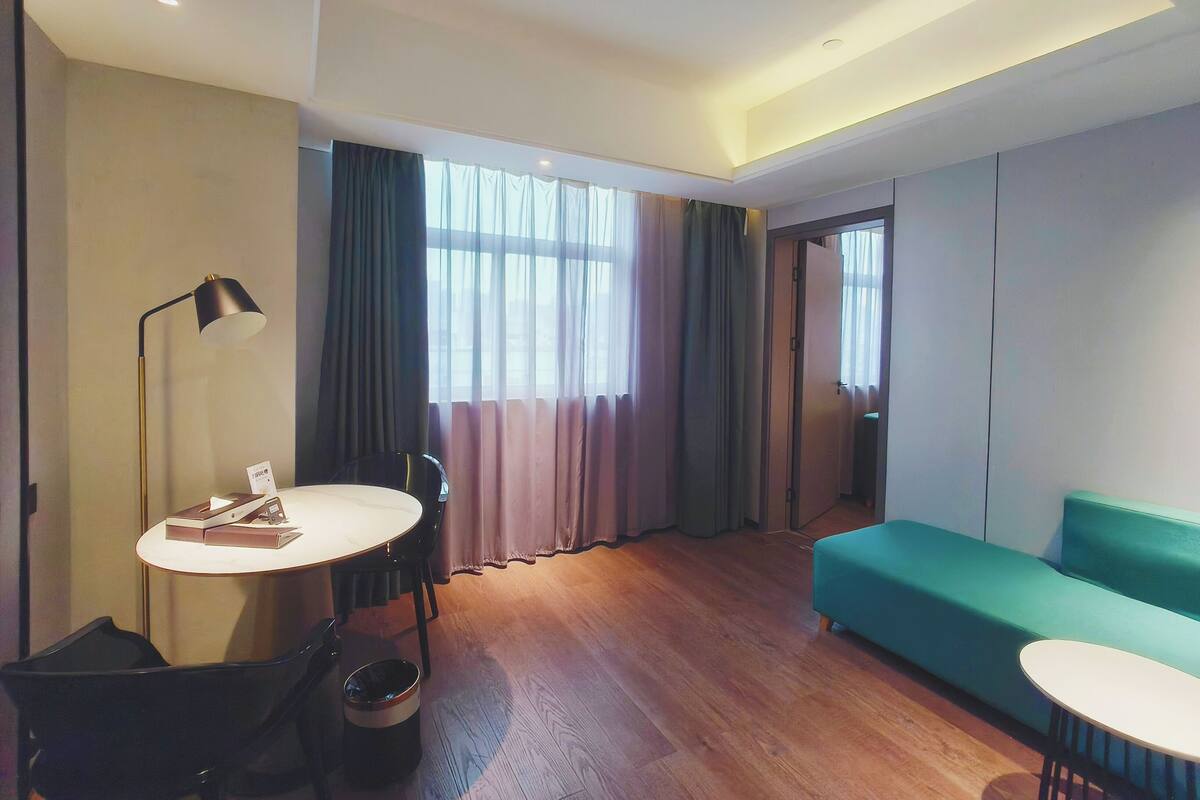 Ein modernes Hotelzimmer mit Holzboden, einem runden Esstisch mit Stühlen, einem grünen Sofa und einem Schreibtisch mit Lampe.
