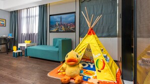 Hotellihuone, jossa on suuri ikkuna, televisio, keltainen ankanmuotoinen pehmolelu, leikkiteltta ja turkoosi sohva.