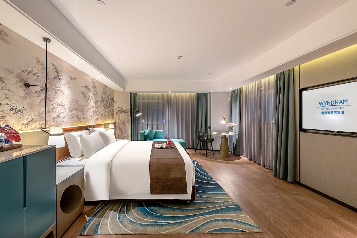 Ein modernes Hotelzimmer mit einem großen Bett, einem Schreibtisch, einem Stuhl, einem kleinen Tisch mit einer Lampe und einem Bildschirm, der den Hotelnamen zeigt.