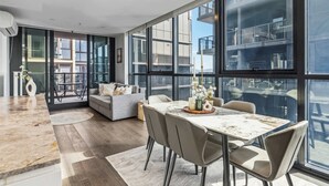 A modern living room with a dining area, large windows, and a view of the city. - Alfred Apartments by Arvia Living (Fortitude Valley)