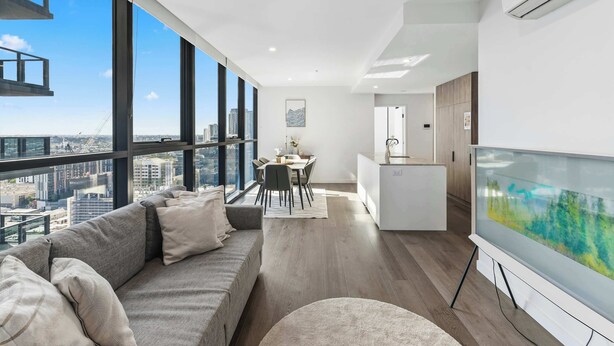 A modern living room with a sofa, a dining area, and a large window offering a city view. - Alfred Apartments by Arvia Living (Fortitude Valley)