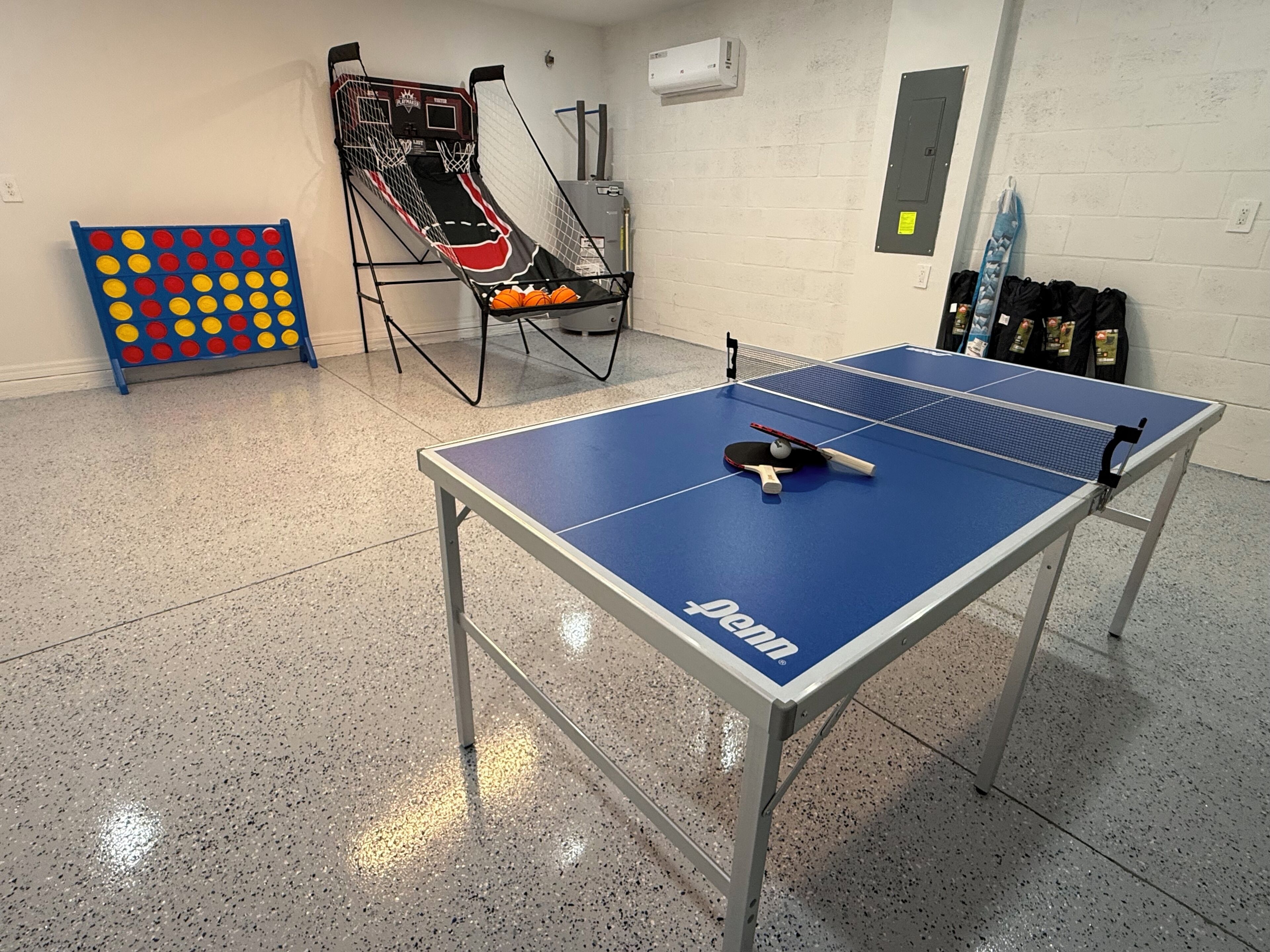 A ping pong table with a net, a basketball hoop, and a dartboard in a room with a tiled floor.