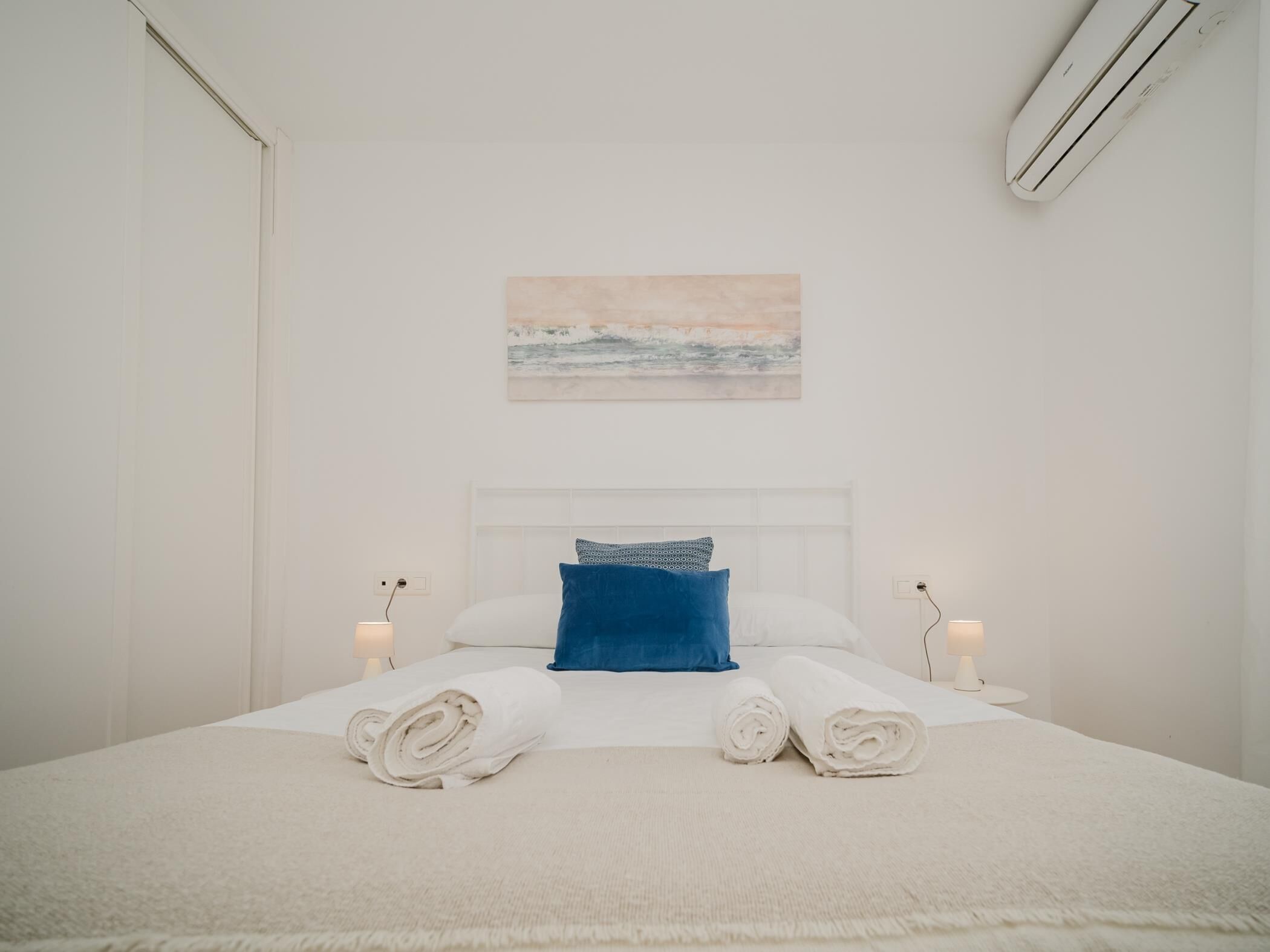 A hotel room with a bed, a headboard, a wall-mounted air conditioner, and a painting.
