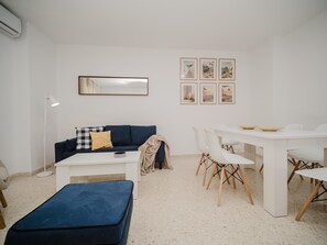 Un salon moderne avec un canapé blanc, un pouf bleu, une table à manger blanche avec des chaises et un grand miroir.