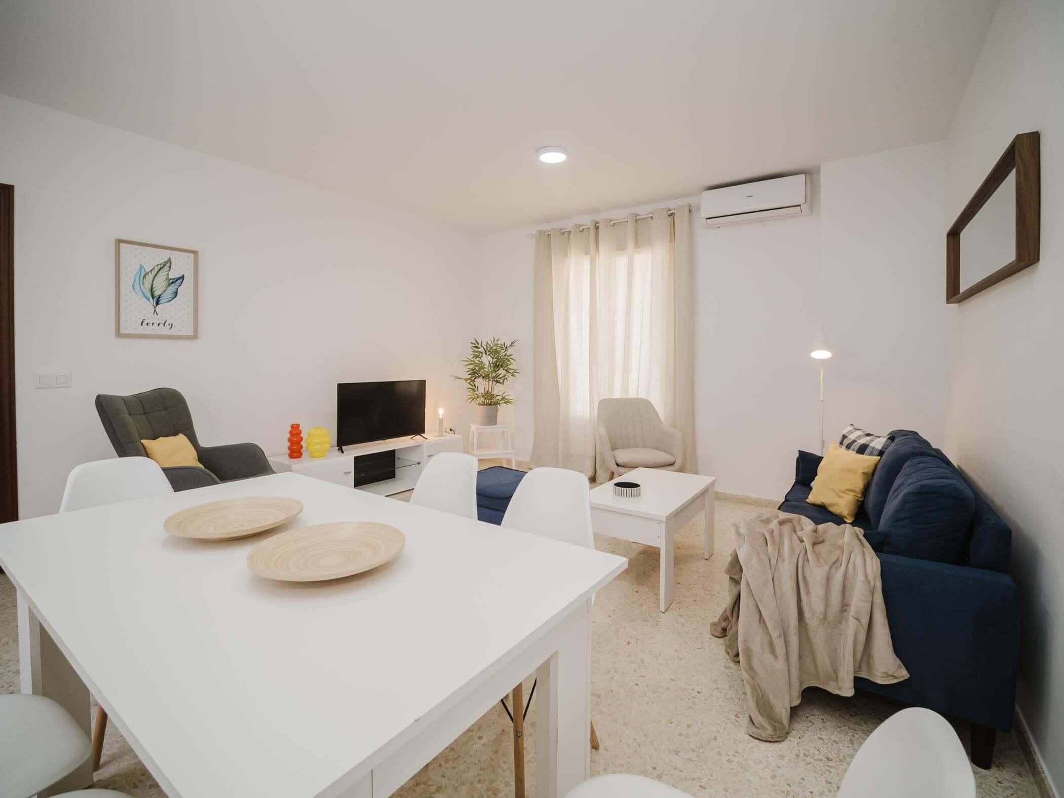 Un salon moderne avec une table à manger blanche, deux canapés, un téléviseur et une petite table sur laquelle est posée une plante.