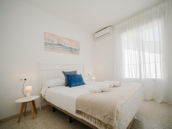 Une chambre à coucher avec un lit, une table de chevet avec une lampe, un climatiseur fixé au mur et une fenêtre avec des rideaux légers.