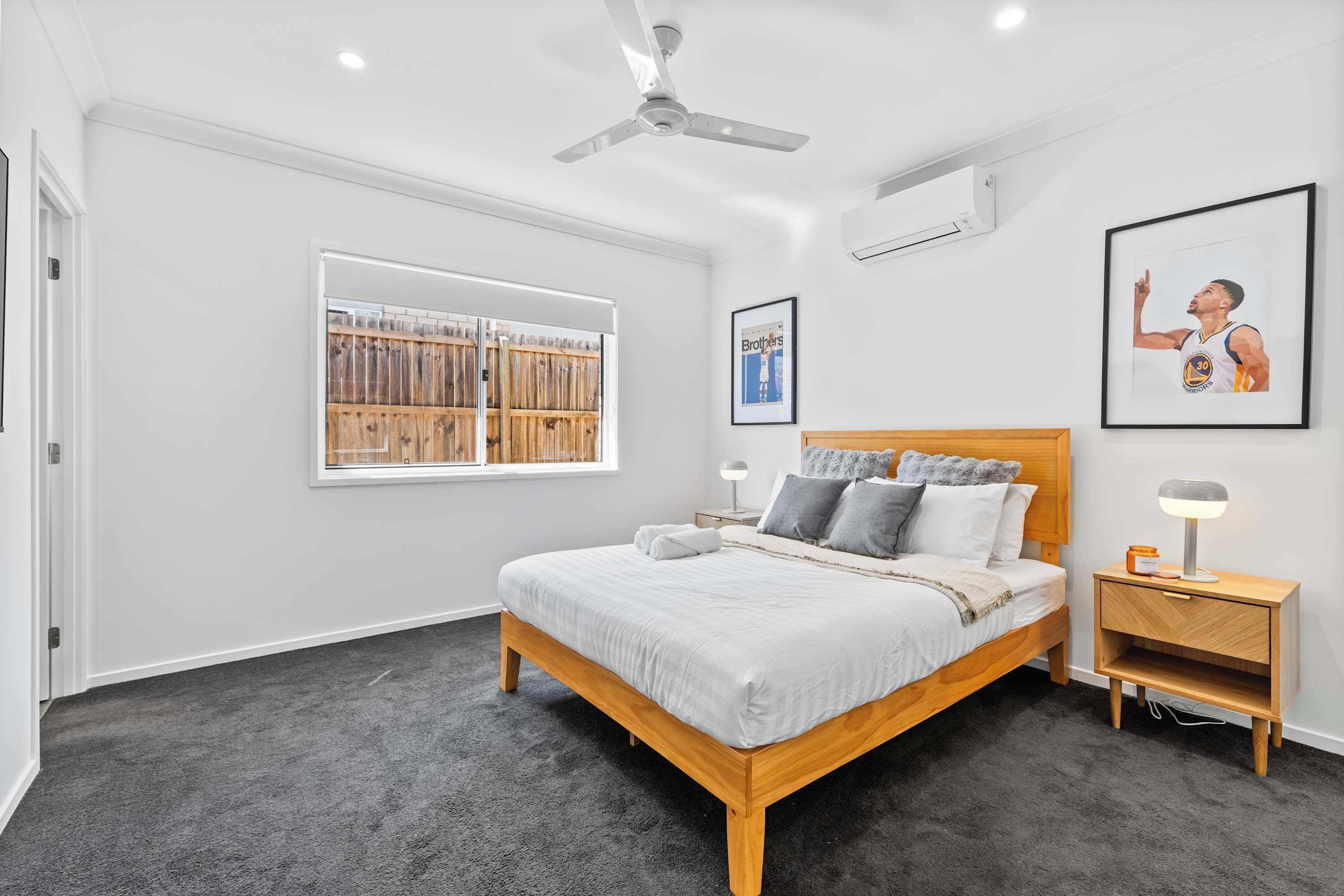 A bedroom with a bed, a nightstand, a ceiling fan, a window with blinds, and framed pictures on the wall.