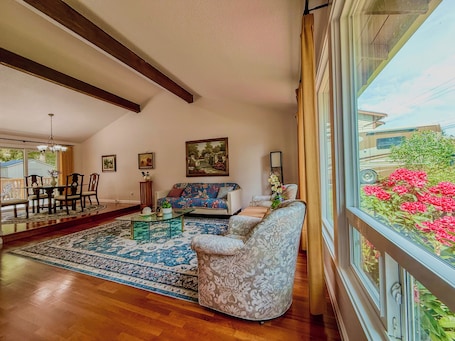 A living room with a sofa, a dining table with chairs, a coffee table, and a large window with a view of greenery and a vehicle.. Entire 3-bedroom charming house in Lake Forest Park