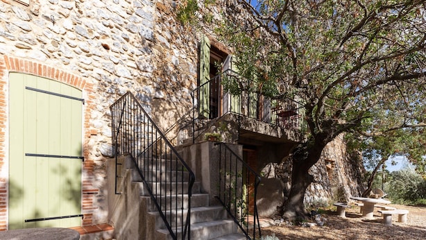 A stone building with a staircase leading to a balcony, surrounded by trees and outdoor seating. - Charming Holiday Home in Tautavel With Balcony (Tautavel)