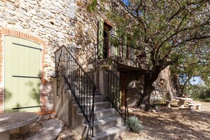 A stone building with a staircase leading to a balcony, surrounded by trees and outdoor seating.