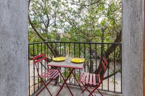 A balcony with a table and chairs, surrounded by a metal railing, overlooking a garden with trees and plants.