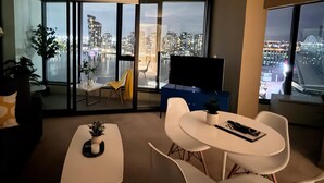 Un salon moderne avec vue sur la ville, un téléviseur à écran plat et un coin repas avec une table et des chaises blanches.