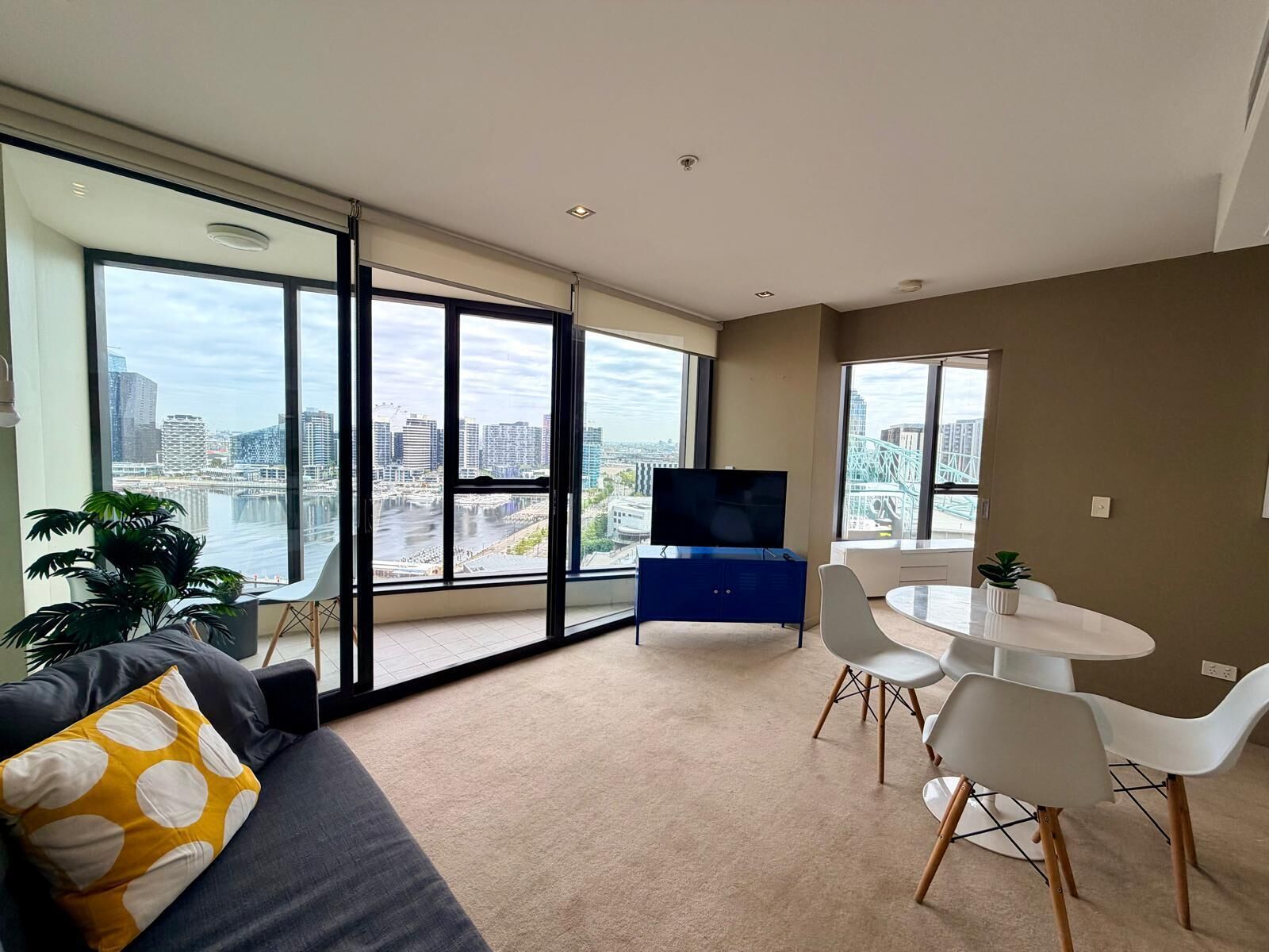 Una sala de estar moderna con ventanas amplias, un televisor de pantalla plana, una zona de comedor con sillas blancas y un sofá con un cojín de lunares.