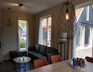 A modern living room with a black leather sofa, a round table, and a coffee table. There are two windows with curtains, a hanging light fixture, and a view of a building outside. - Modern Villa With Garden (Almen)