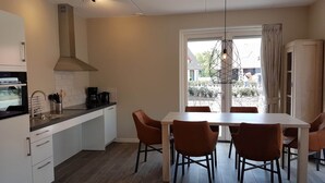 A modern kitchen with a dining area, featuring a large window, a hanging light fixture, and a built-in oven. - Modern Villa With Garden (Almen)