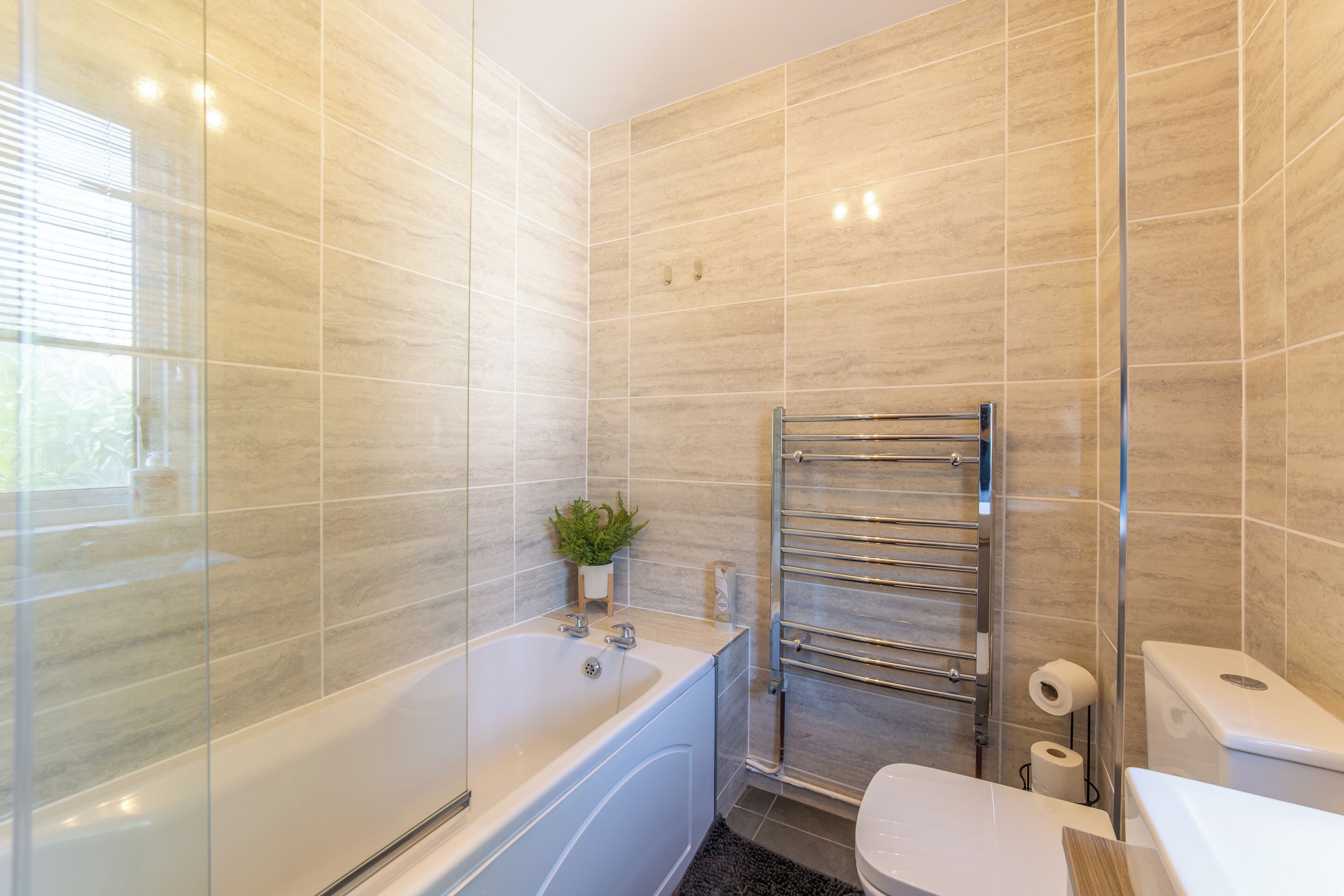 A modern bathroom with a glass-enclosed shower, a white bathtub, a glass shower door, a glass-enclosed toilet, and a small potted plant on the sink.