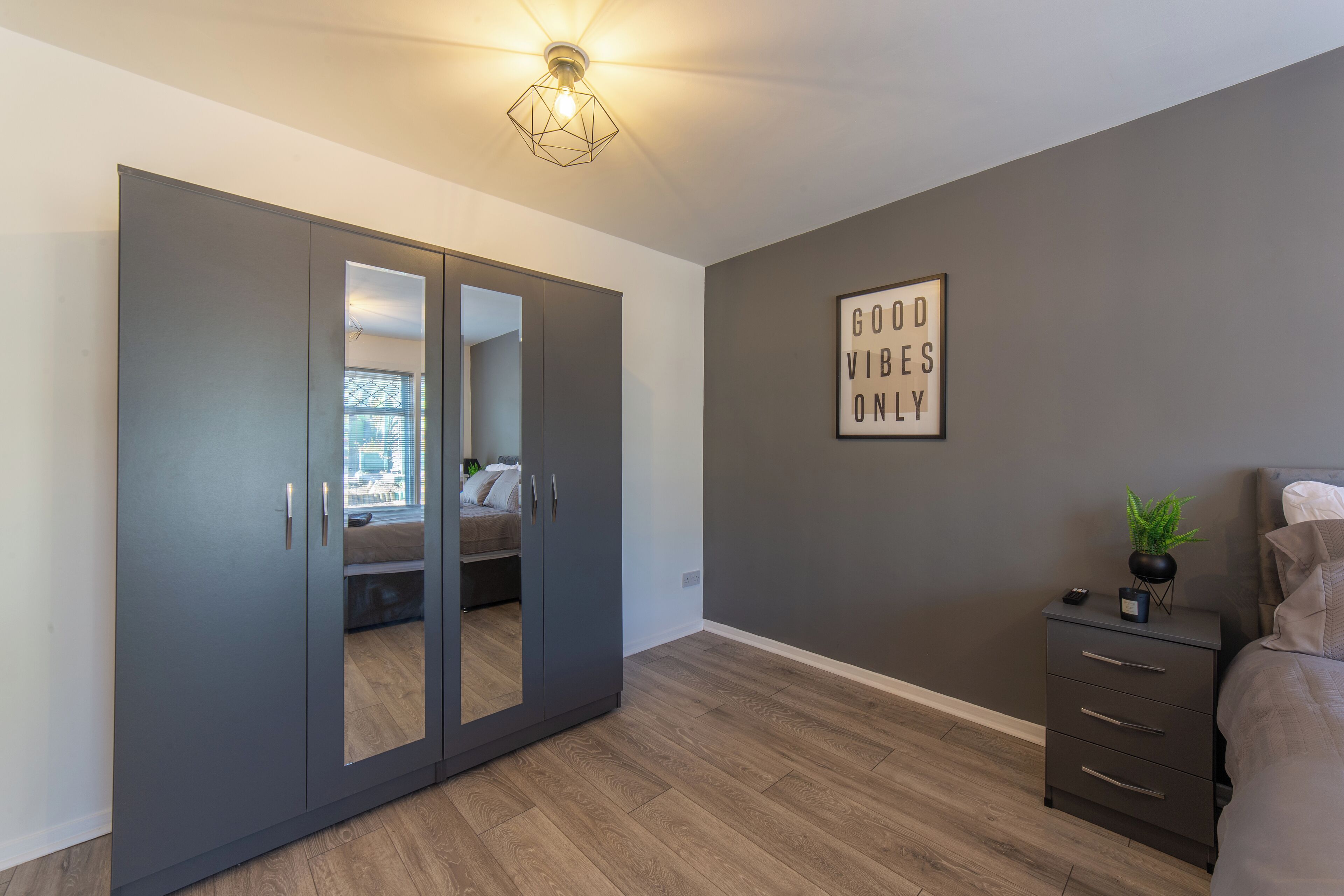 A bedroom with a wardrobe, a bedside table with a plant, and a motivational quote on the wall.