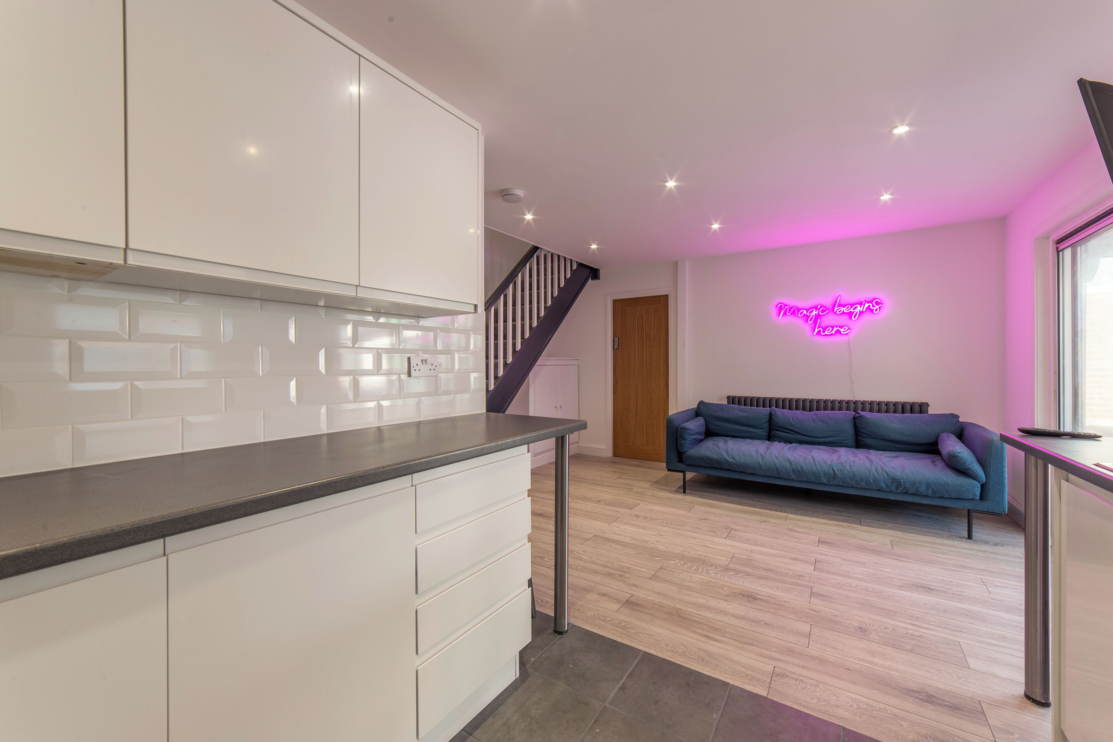 A modern kitchen with white cabinets and a grey countertop, a staircase leading to a loft area, and a neon sign on the wall.