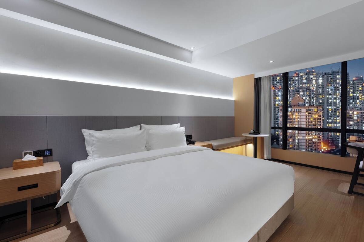 A modern hotel room with a large bed, bedside table, and a city view through large windows.
