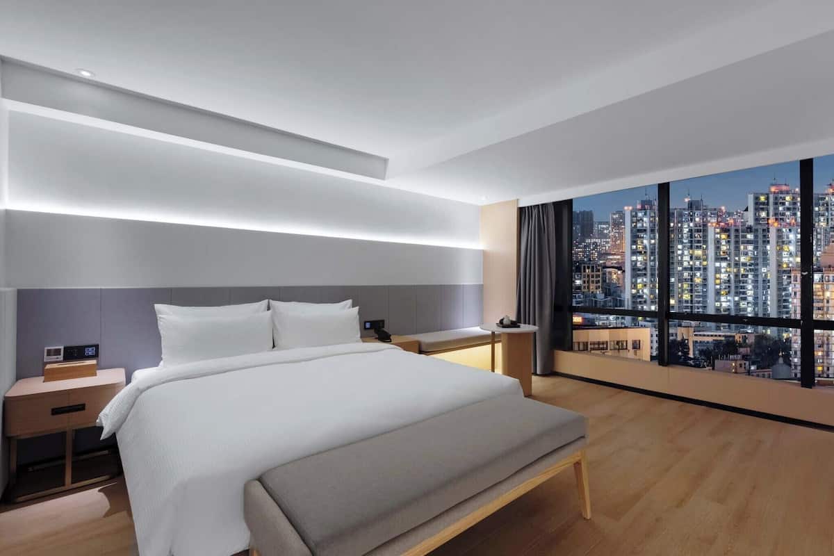 A modern hotel room with a large bed, bedside tables, a nightstand, and a view of a cityscape through large windows.