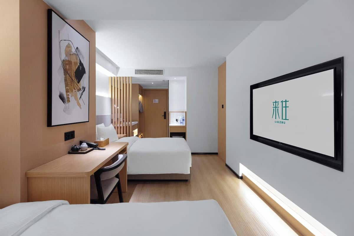 A hotel room with two beds, a desk, a chair, a bedside table, a lamp, and a framed artwork on the wall.
