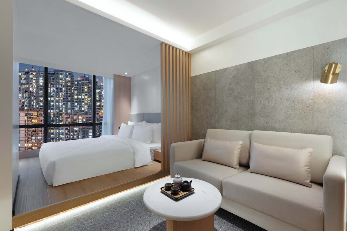 A modern hotel room with a large bed, a sofa, a round coffee table with a tray of tea and cups, and a cityscape view through large windows.