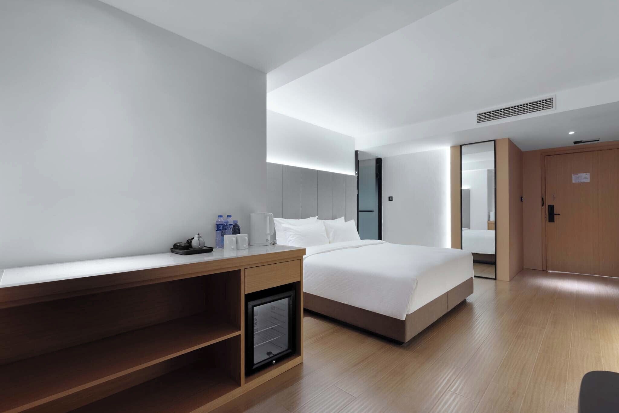 A modern hotel room with a wooden desk, a bed with white linens, a built-in mini-fridge, and a large mirror on the wall.