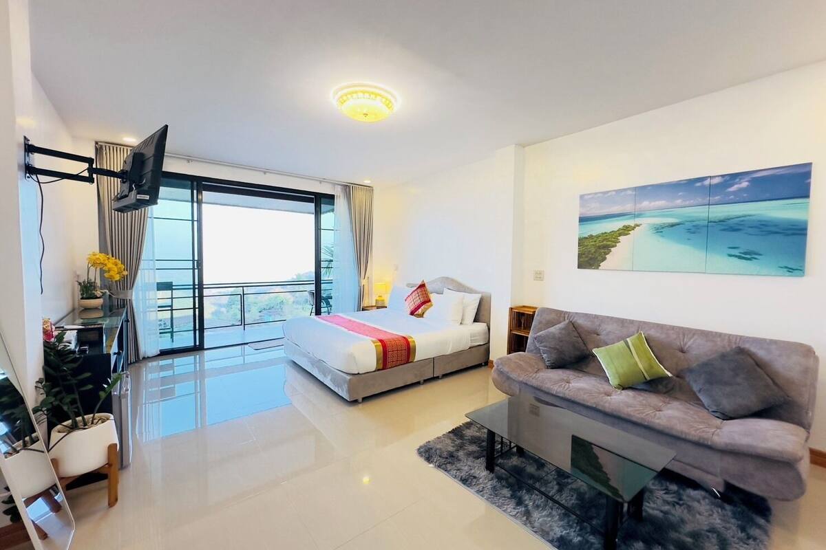 A modern hotel room with a balcony, a large bed, a sofa, and a painting of a beach scene.