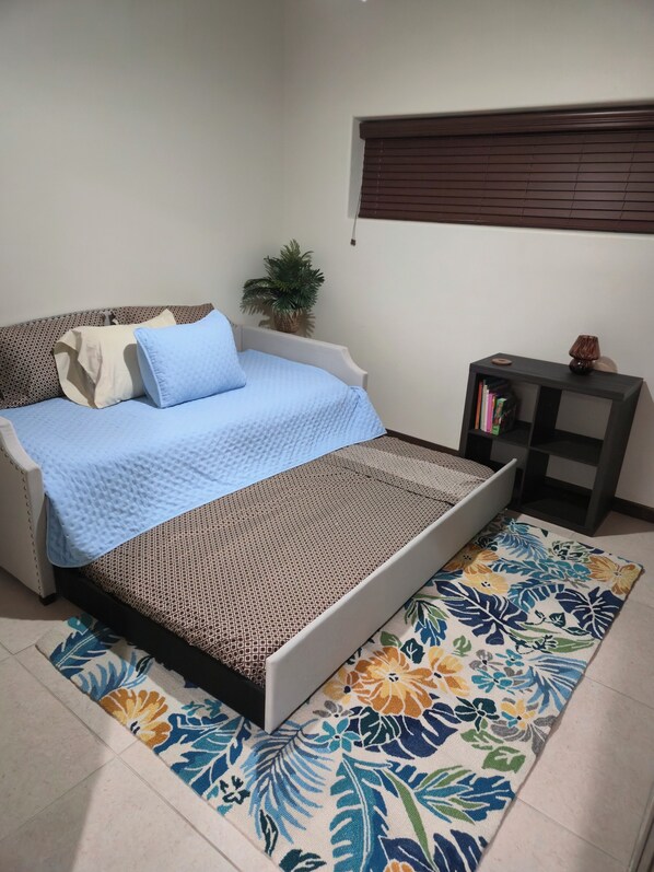 A bedroom with a bed, a small side table, a potted plant, and a window with blinds.