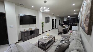 Uma sala de estar moderna com televisão de tela plana, móveis de madeira escura, uma mesa de centro e um espaço de refeições com cadeiras e mesa.