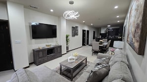 A modern living room with a flat-screen TV, dark wood furniture, a coffee table, and a dining area with chairs and a table. - Spacious 3Bedroom Apartment in Gated Community Prime Location, 1Mi from Shopping (Mission)