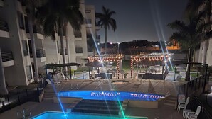 Piscina com um elemento aquático, rodeada por palmeiras e um edifício com área de estar ao ar livre.