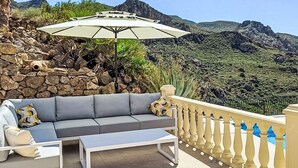 Ein Balkon mit einer grauen Couch, einem kleinen Tisch und einem Sonnenschirm. Der Balkon bietet Blick auf eine felsige Landschaft mit viel Grün und Bergen im Hintergrund.