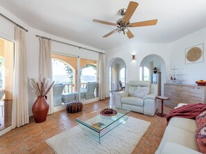 A spacious living room with a glass coffee table, a ceiling fan, a sofa, and a view through large glass doors.
