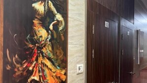 A corridor with a large painting of a woman in a colorful dress on the wall, wooden paneling, and a series of doors with numbers. - metroview rooms & celebration (New Delhi)