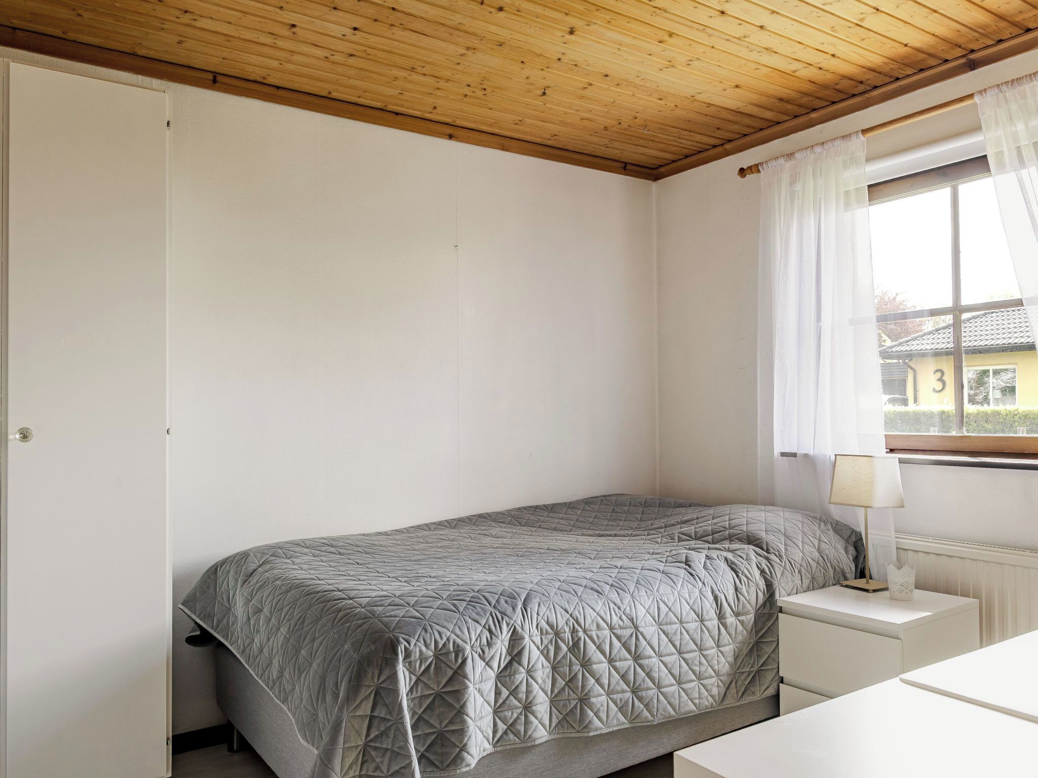 Une chambre avec un lit simple, une table de chevet, une fenêtre avec des rideaux et un plafond en bois.