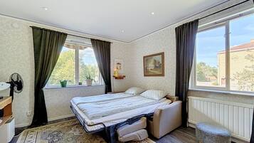 Une chambre à coucher comprenant un lit, une table de chevet, une chaise, une fenêtre avec des rideaux, un radiateur et un petit tabouret.