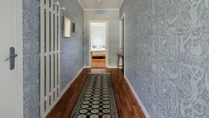 A hallway with patterned wallpaper, a wooden floor, a black and white patterned rug, a bed with a white headboard, and a hanging light fixture.