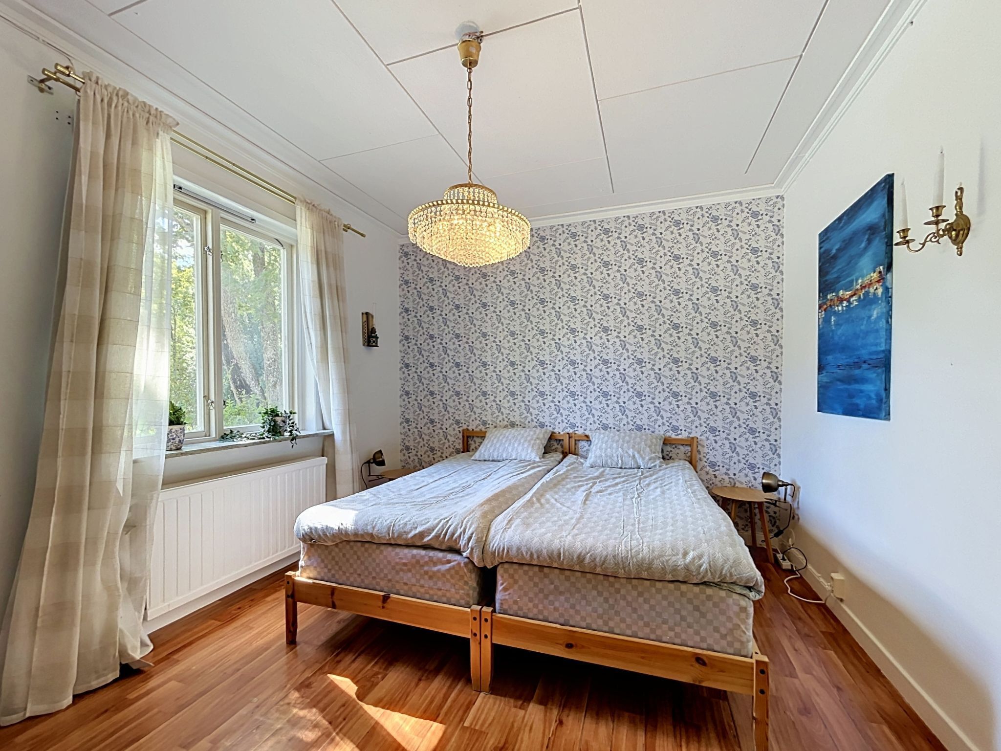 A bedroom with a wooden bed, a window with curtains, a patterned wallpaper, and a painting on the wall.