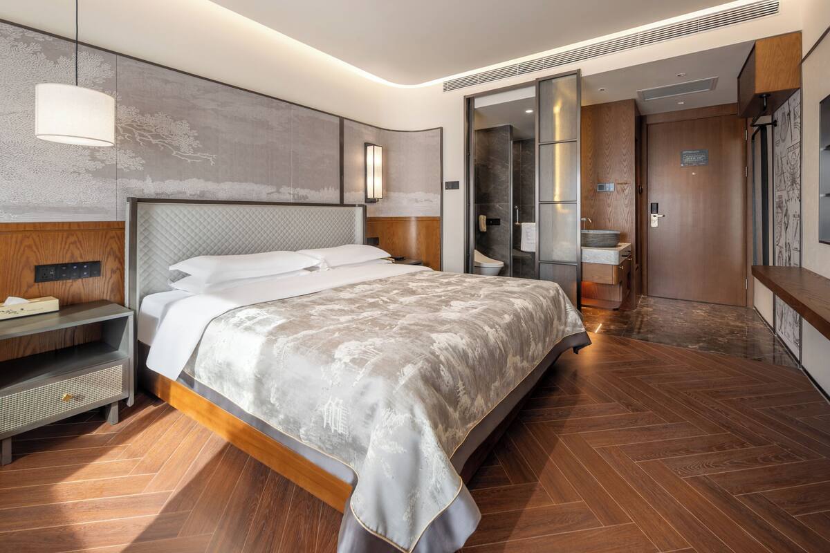 A modern hotel room with a large bed, a bedside table, a built-in wardrobe, and a bathroom with a shower and toilet.