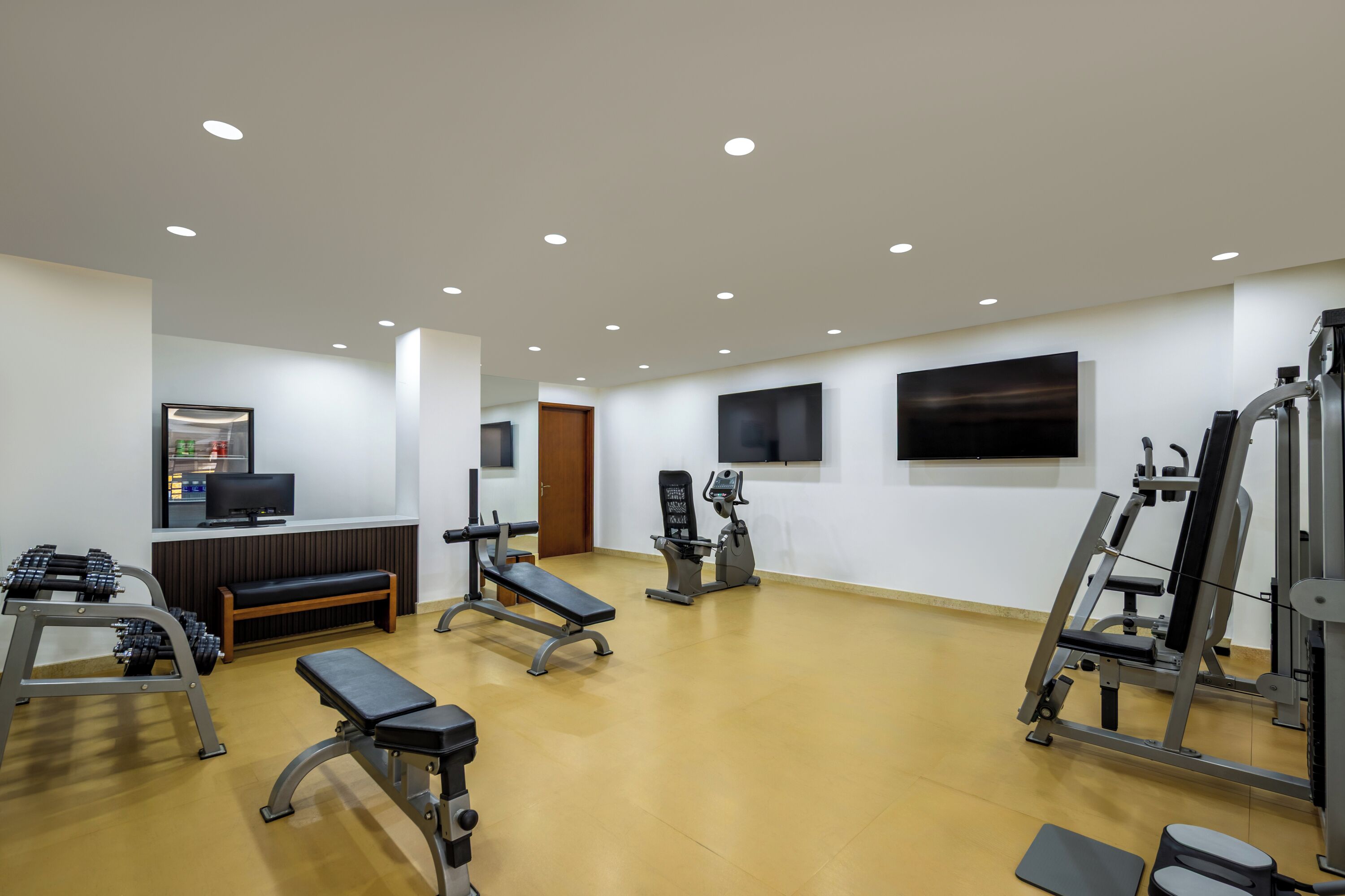 Uma academia com equipamentos de exercícios, um banco, uma televisão e uma TV fixada na parede.