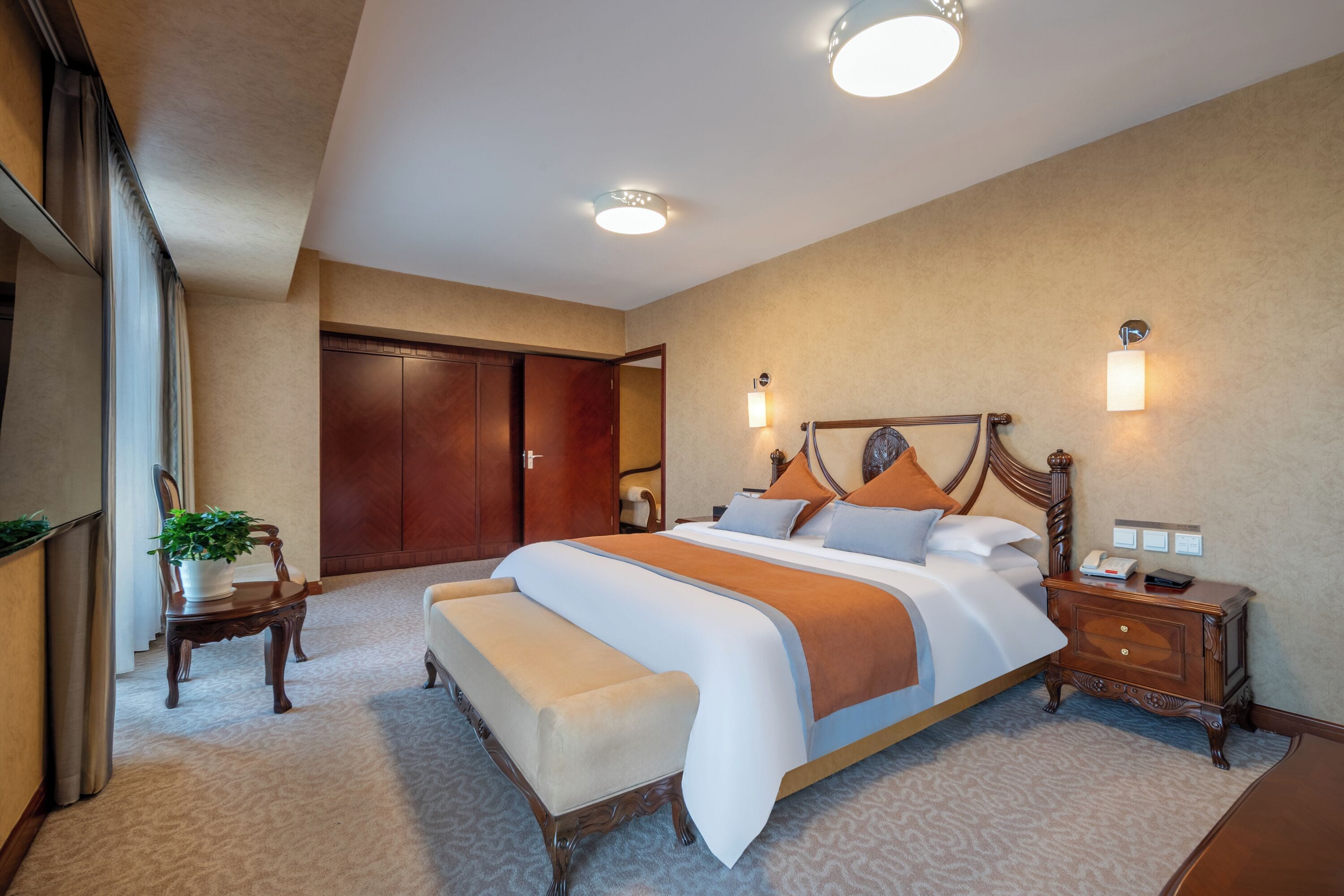 A hotel room with a large bed, a wooden nightstand, a bench, a potted plant, and a wardrobe.