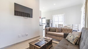 Una sala de estar moderna con un sofá, una mesa de centro con libros, un televisor de pantalla plana en la pared y una cocina al fondo.