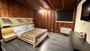 A bedroom with a wooden bed, a bedside table, a dresser, a TV mounted on the wall, and a window with blinds.