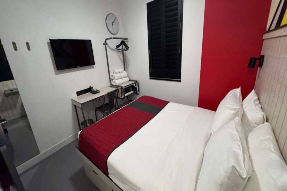 A bedroom with a bed, a TV, a desk with a chair, a towel rack, and a window with shutters.