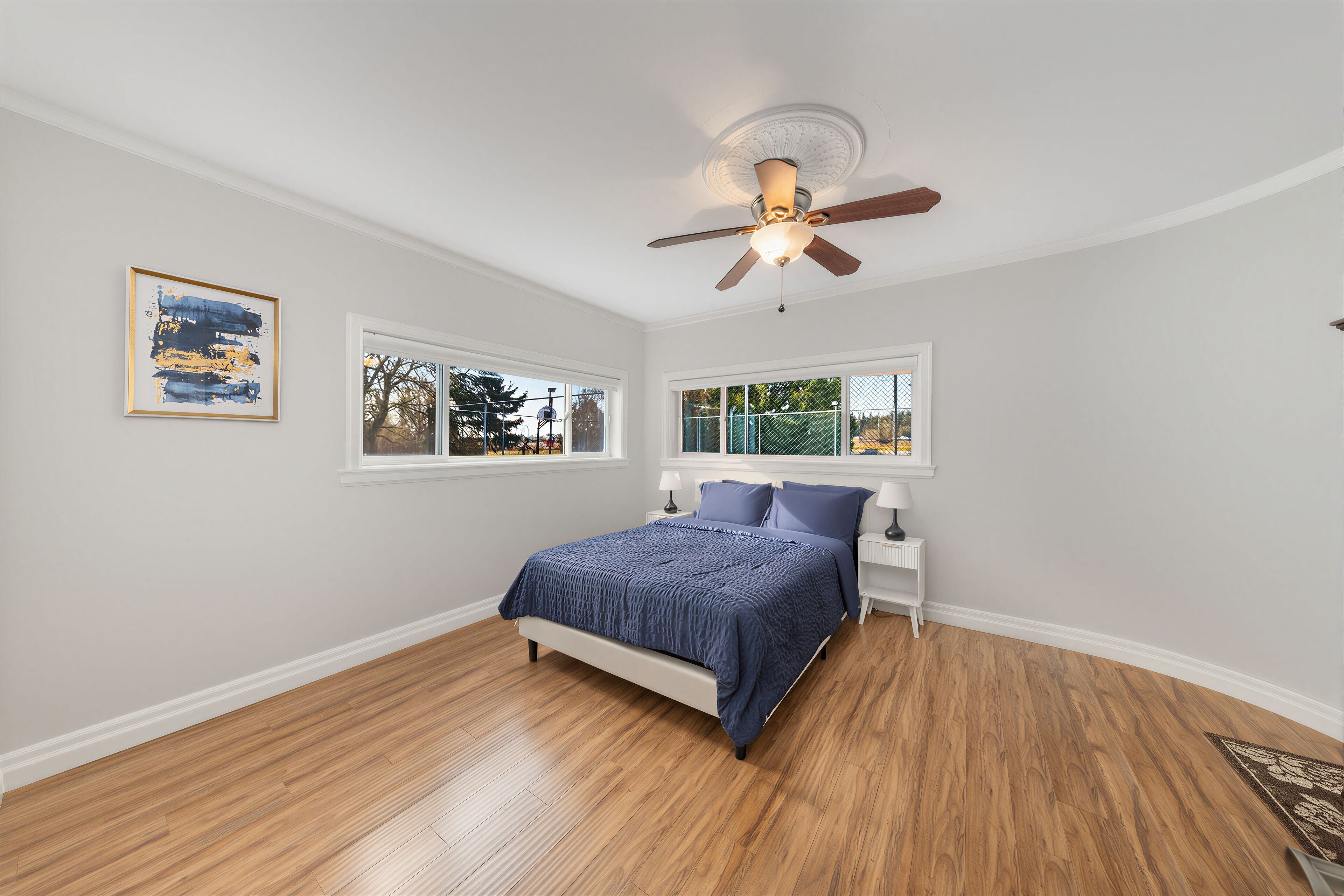 A bedroom with a bed, two windows, a ceiling fan, and a framed picture on the wall.