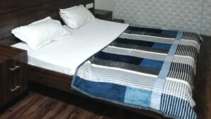 Een enkel bed met een houten hoofdbord, wit beddengoed en een gestreept deken. Boven het bed is een plafondventilator te zien, en een airconditioningunit is aan de muur bevestigd.