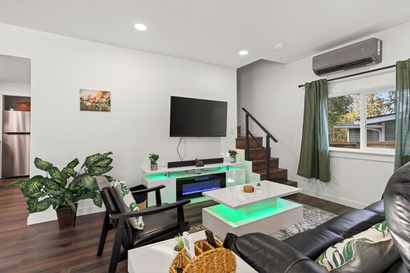A modern living room with a flat-screen TV, a fireplace, a staircase, and a large window with curtains. - Emerald City Jungle Oasis – 10 Min to Downtown Seattle (Seattle)