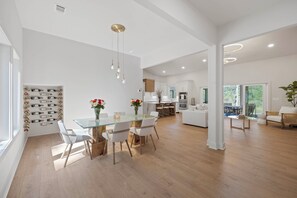 A spacious living area with a glass dining table, white chairs, and a modern kitchen in the background. - 'The Emerald Cove Residence' Lake Oconee Luxury! (Eatonton)