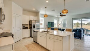 A modern kitchen with white cabinetry, a central island, and a built-in dishwasher. There is a ceiling fan and pendant lighting. - Golfers Dream (Golf Included) – IBIS Landing Golf (Lehigh Acres)